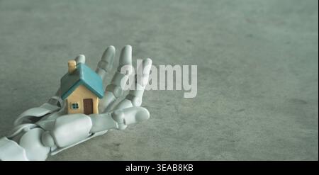 A white futuristic robotic hand gently cradling a small toy house against a plain grey background. Stock Photo