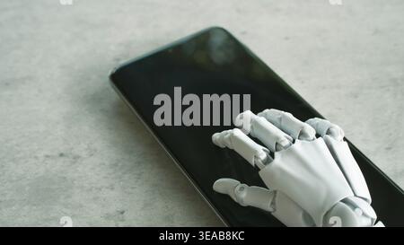 A white prosthetic robot hand interacts with a modern black smartphone resting on a grey stone surface. Stock Photo