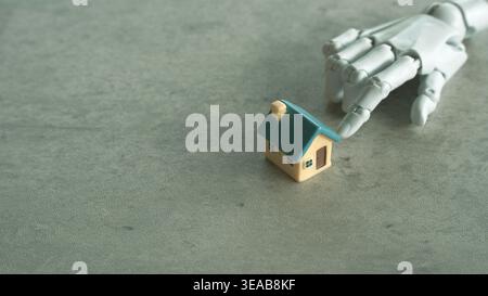A white robotic hand reaches toward a miniature house model on a concrete surface. Stock Photo