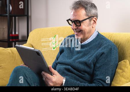 An elderly man seated on a yellow couch, utilizing technology for leisure, signified by digital task checkmarks. Stock Photo