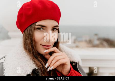 Woman red beret fashion stylish woman wearing a shearling jacket and red beret poses outdoors near the ocean on a cloudy day Stock Photo