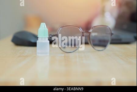 Clear lubricating eye drop bottle placed on a wooden table next to a pair of glasses with laptop, symbolizing relief for dry eyes. Stock Photo