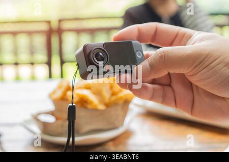 Cropped view close-up of unrecognizable photographer holding mini black action camera while taking pictures of food on table, selective focus. Stock Photo