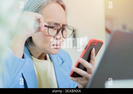 Mature Asian businesswoman with bad sight trying to read message on screen of mobile phone. Eye problem concept. Stock Photo