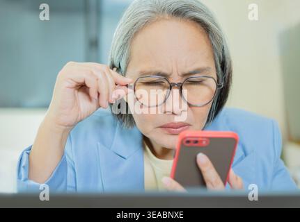Mature woman wearing blue suit holding smartphone at the office, struggling to focus while reading a message. Concept of eye strain, vision problems, Stock Photo