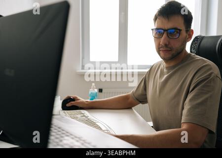 Focused adult male professional in glasses sits at desk, concentrating on computer screen. Natural light from window illuminates home office workspace Stock Photo
