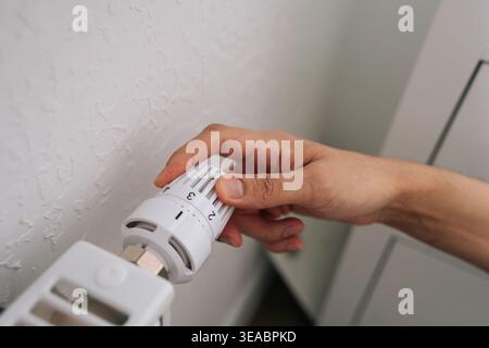 Person hand adjusting white thermostat knob on modern radiator. Close-up view with shallow depth of field. Concept of energy saving, heating costs Stock Photo