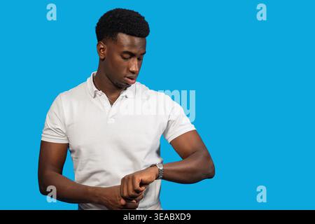 Young black man checking his watch, focusing on punctuality and waiting on blue background Stock Photo