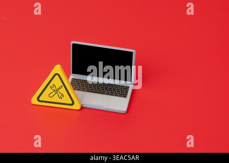 A miniature laptop next to a warning sign displaying a wrench and screwdriver icon. Minimalist concept representing IT support, computer repair Stock Photo