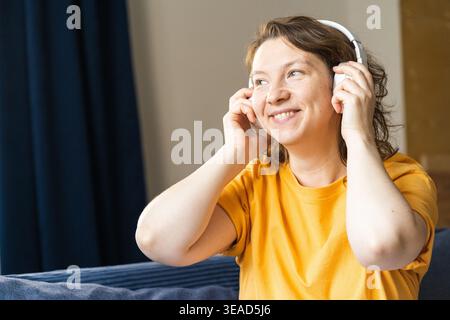Woman wearing a yellow t-shirt and wireless headphones, smiling while listening to music and relaxing at home. Portrait of a young joyful woman Stock Photo