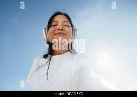 Young woman listening to music with over-ear headphones outdoors, eyes closed and smiling while enjoying sunlight and calm. Image conveys relaxation, Stock Photo