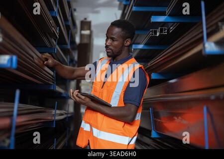African American worker with a reflective safety vest using a digital tablet while taking inventory in a metalworking facility warehouse. Stock Photo