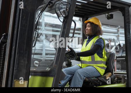 Portrait of a smiling female worker wearing protective gear, a yellow hard hat and a safety vest, driving a forklift truck in a warehouse. Stock Photo