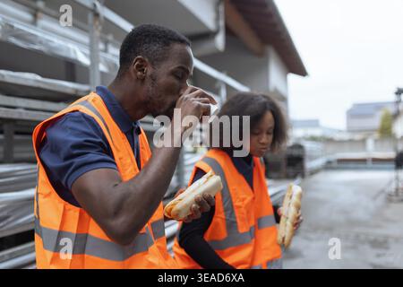 Two coworkers wearing reflective vests having fun during a lunch break, eating sandwiches outdoors at an industrial site. Stock Photo