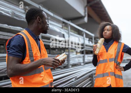 Two coworkers wearing reflective vests having fun during a lunch break, eating sandwiches outdoors at an industrial site. Stock Photo