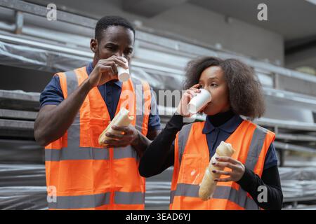 Two colleagues, warehouse workers having a lunch break, enjoying sandwiches and relaxed chat with a smile and coffee after a hard work. Stock Photo