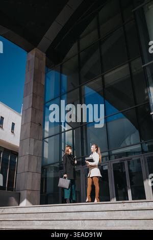 Two businesswomen talking on steps outside a modern glass office building during daytime Stock Photo