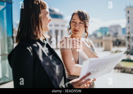 Two businesswomen discussing documents outside an office building during a daytime city meeting Stock Photo
