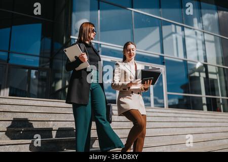 Two businesswomen walking down steps outside a modern office building discussing documents Stock Photo