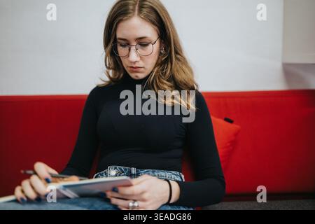 Young student studying with notebook on red couch, focused woman wearing glasses and jeans Stock Photo