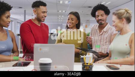 Discussing team in work clothes pointing at silver laptop in office, showing tablet overlays Stock Photo