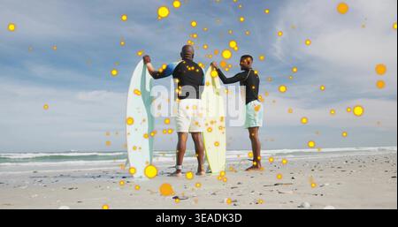 Holding boards, two men in rash guards and shorts on shoreline preparing to surf, orange particles Stock Photo