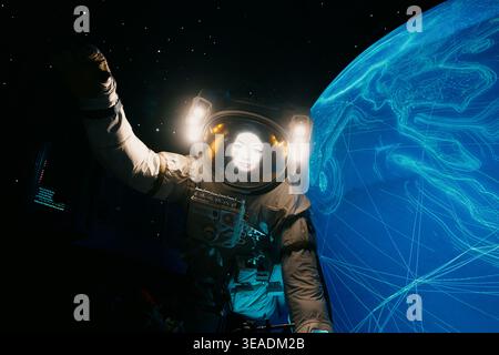 13 January 2026, Dubai Expo City, UAE: Female astronaut wearing a space suit, helmet showing a woman's face, floating next to a digital blue Earth in Stock Photo