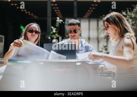 Three young business people collaborating over a laptop and documents at an outdoor cafe meeting Stock Photo