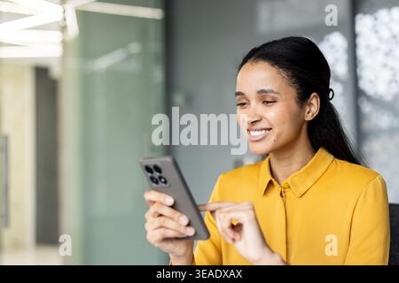 Young mixed-race business woman smiling, using a mobile phone, and feeling happy while working in a modern office creating a positive work environment Stock Photo