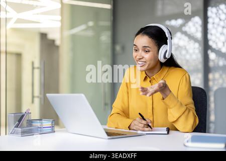 Young woman working from home, smiling and engaged in a virtual meeting at a white desk, taking notes while using laptop and headphones for online communication and learning Stock Photo