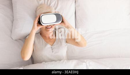 Senior woman lying on bed and using virtual reality headset Stock Photo