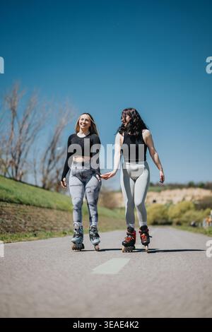 Two young women rollerblading together on a sunny suburban path holding hands and smiling Stock Photo
