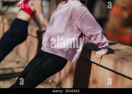 Two women doing triceps dips on a stone wall during an outdoor fitness workout Stock Photo