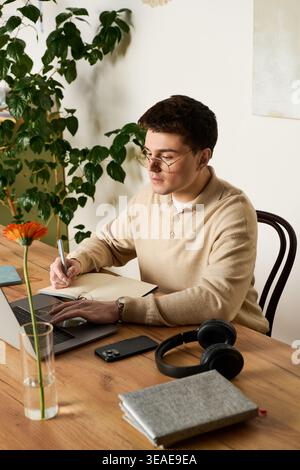 A young man sits at a wooden table, writing notes while surrounded by greenery and calm ambiance. Stock Photo