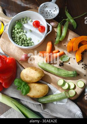 Assorted organic vegetables and vintage utensils on wooden board in sunlit countryside kitchen. High-angle shot captures sliced pumpkin carrots Stock Photo
