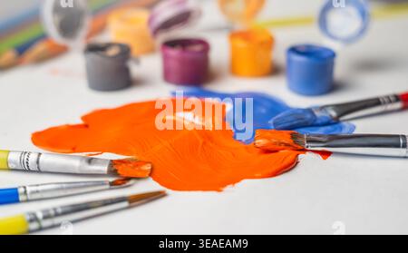 Two brushes rest on vibrant orange and blue paint on a work surface surrounded by paint containers in the afternoon light. Stock Photo