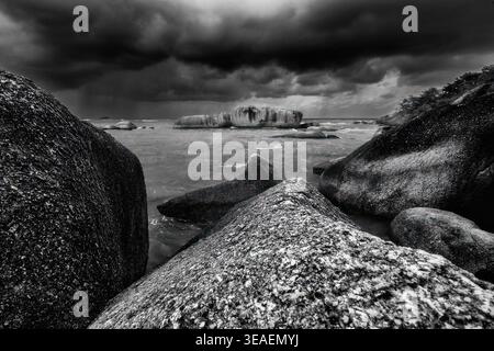 Dramatic black and white seascape with large coastal boulders leading into a stormy horizon. The dark clouds above create an ominous, cinematic tensio Stock Photo
