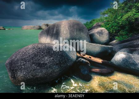 The photograph captures a dramatic scene of a tropical coastline with huge granite boulders surrounded by green water and an ominous dark sky. In the Stock Photo