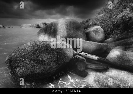 The black and white photograph captures a dramatic coastline with massive boulders, wild clouds and a calm sea in the background. The image combines t Stock Photo