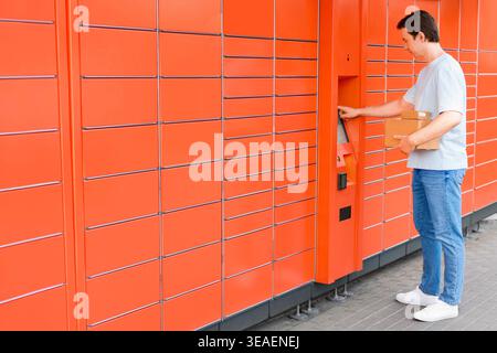 Man with cardboard boxes uses the touch screen of a large orange automated parcel locker outdoors. Self-service delivery, returning online orders, and Stock Photo
