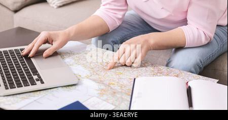 Person Using Laptop While Looking at Map in Living Room With Notebook Open on Table Stock Photo