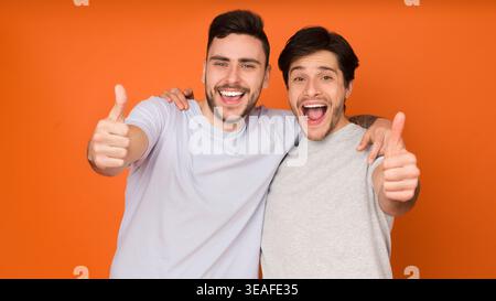 Two Friends Smile and Pose Together in Front of Bright Orange Background With Thumbs Up Stock Photo