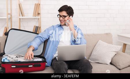 Man on Couch Packs Suitcase While Talking on Phone and Using Computer in Modern Living Room Stock Photo