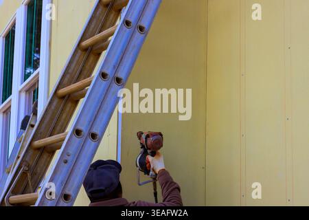Worker uses pneumatic nail gun to installing siding outside of building while standing on ladder Stock Photo