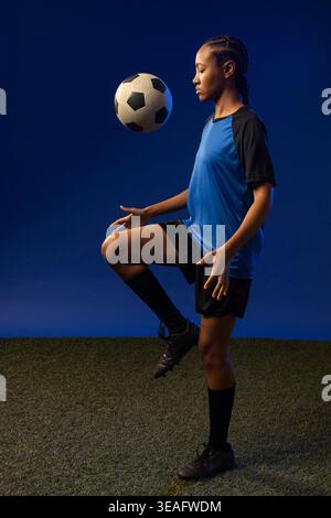 African female athlete balancing classic paneled soccer ball near raised knee on turf in blue kit Stock Photo