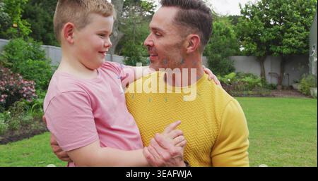 Holding father in yellow sweater cradling son in pink T-shirt in residential backyard, with lawn Stock Photo