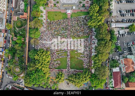 Aerial view of a large crowd of people gathered for communal prayer in a green city square surrounded by lush trees and urban streets in Klaten, Jawa Stock Photo