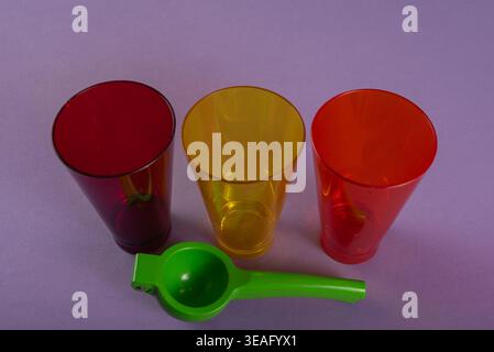 Still life featuring transparent plastic cups (red, yellow, orange) plus a lime squeezer on a purple background. Soft studio lighting emphasizes vivid Stock Photo