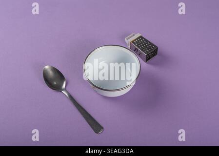 Still life with metal spoon, white bowl, and small grater arranged on solid purple background, studio lit. Promotes kitchenware, food preparation, and Stock Photo