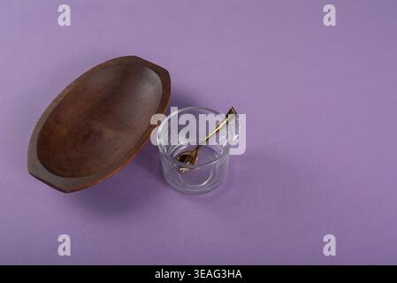 Still life features an empty wooden bowl, glass cup, and golden spoon. Soft light enhances smooth textures. Background: solid violet. Useful for food, Stock Photo
